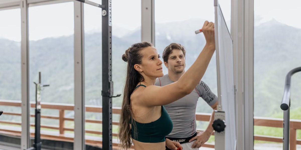 Female fitness coach writes on whiteboard as a male athlete watches in a gym.