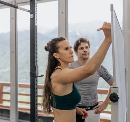 Female fitness coach writes on whiteboard as a male athlete watches in a gym.